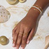 A hand adorned with the Celia Micro Freshwater Pearl Bracelet in Gold, along with gold and white rings, rests on a white surface scattered with gold glitter, ornaments, and a satin ribbon.