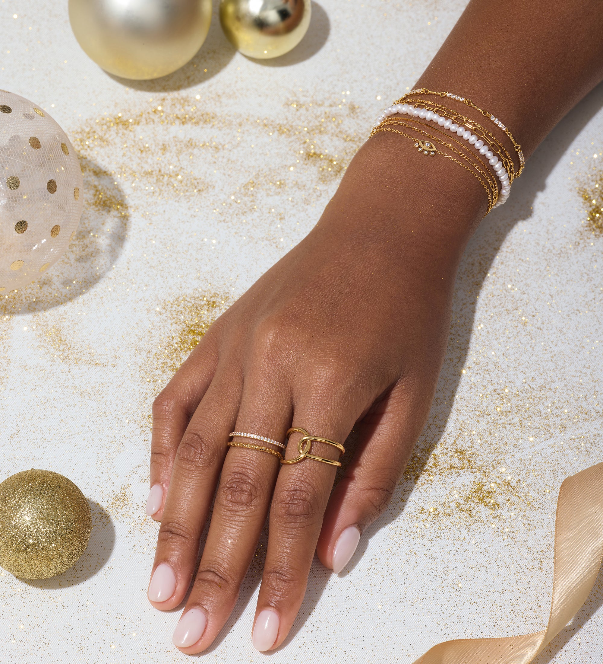 A hand adorned with the Celia Micro Freshwater Pearl Bracelet in Gold, along with gold and white rings, rests on a white surface scattered with gold glitter, ornaments, and a satin ribbon.
