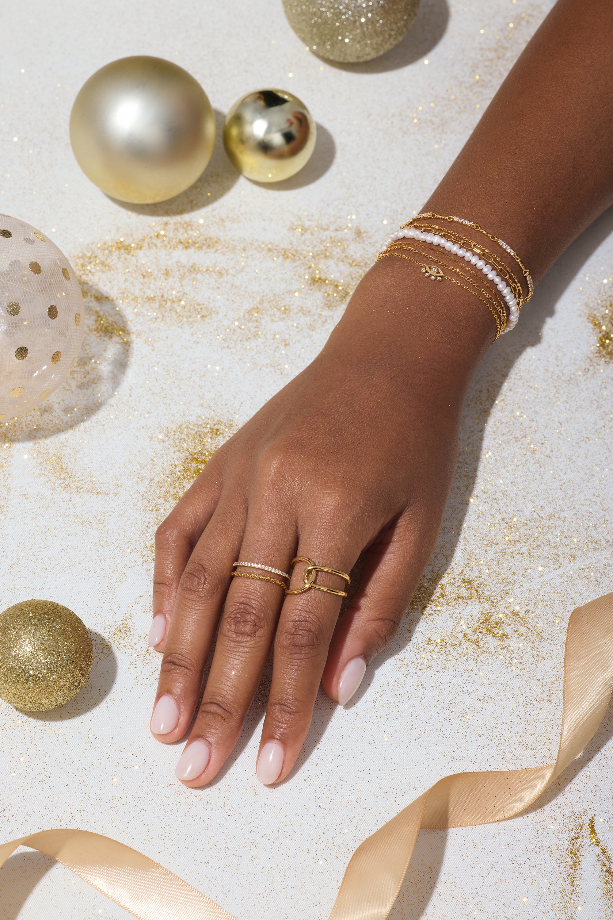A hand adorned with the Celia Micro Freshwater Pearl Bracelet in Gold, along with gold and white rings, rests on a white surface scattered with gold glitter, ornaments, and a satin ribbon.