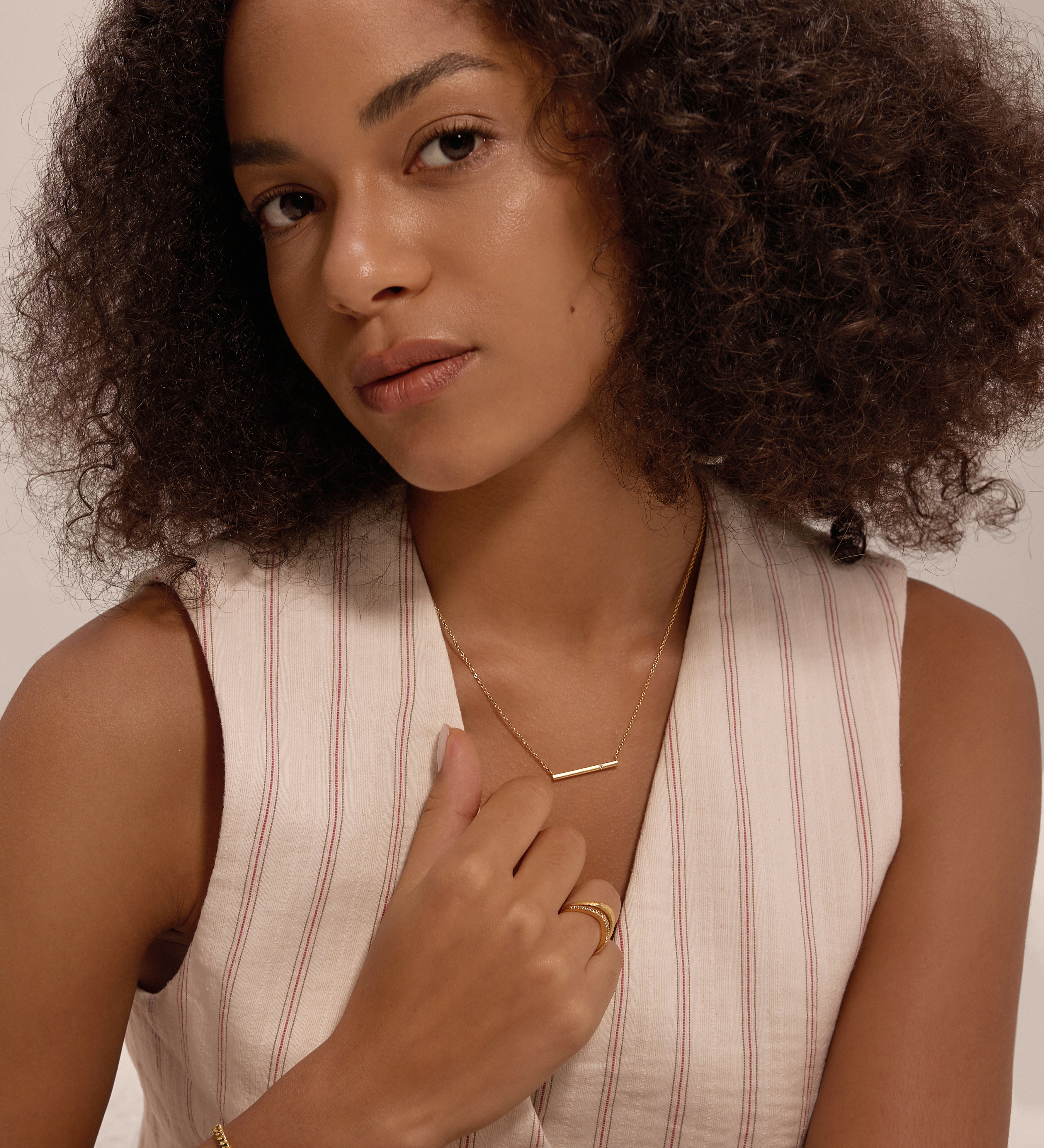 A woman with curly hair wears a sleeveless, light-striped top and minimalist jewellery, including the Regina Horizontal Bar Pendant Necklace in Gold, along with cubic zirconia rings, posing against a neutral background.