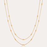 The Simona Double Chain Stacking Necklace in Gold features two delicate chains with evenly spaced oval and round bead accents, making it a minimalist piece thats perfect for layering or wearing solo against a light background.
