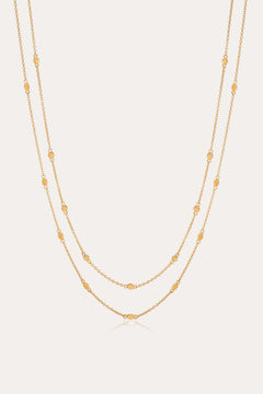 The Simona Double Chain Stacking Necklace in Gold features two delicate chains with evenly spaced oval and round bead accents, making it a minimalist piece thats perfect for layering or wearing solo against a light background.