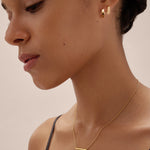A woman with smooth skin and dark hair tied back wears the Juliet Angular Huggies in Gold, a delicate gold bar necklace, and a black strap top, photographed in soft lighting against a neutral background.
