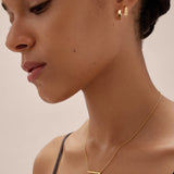 A woman with smooth skin and dark hair tied back wears the Juliet Angular Huggies in Gold, a delicate gold bar necklace, and a black strap top, photographed in soft lighting against a neutral background.