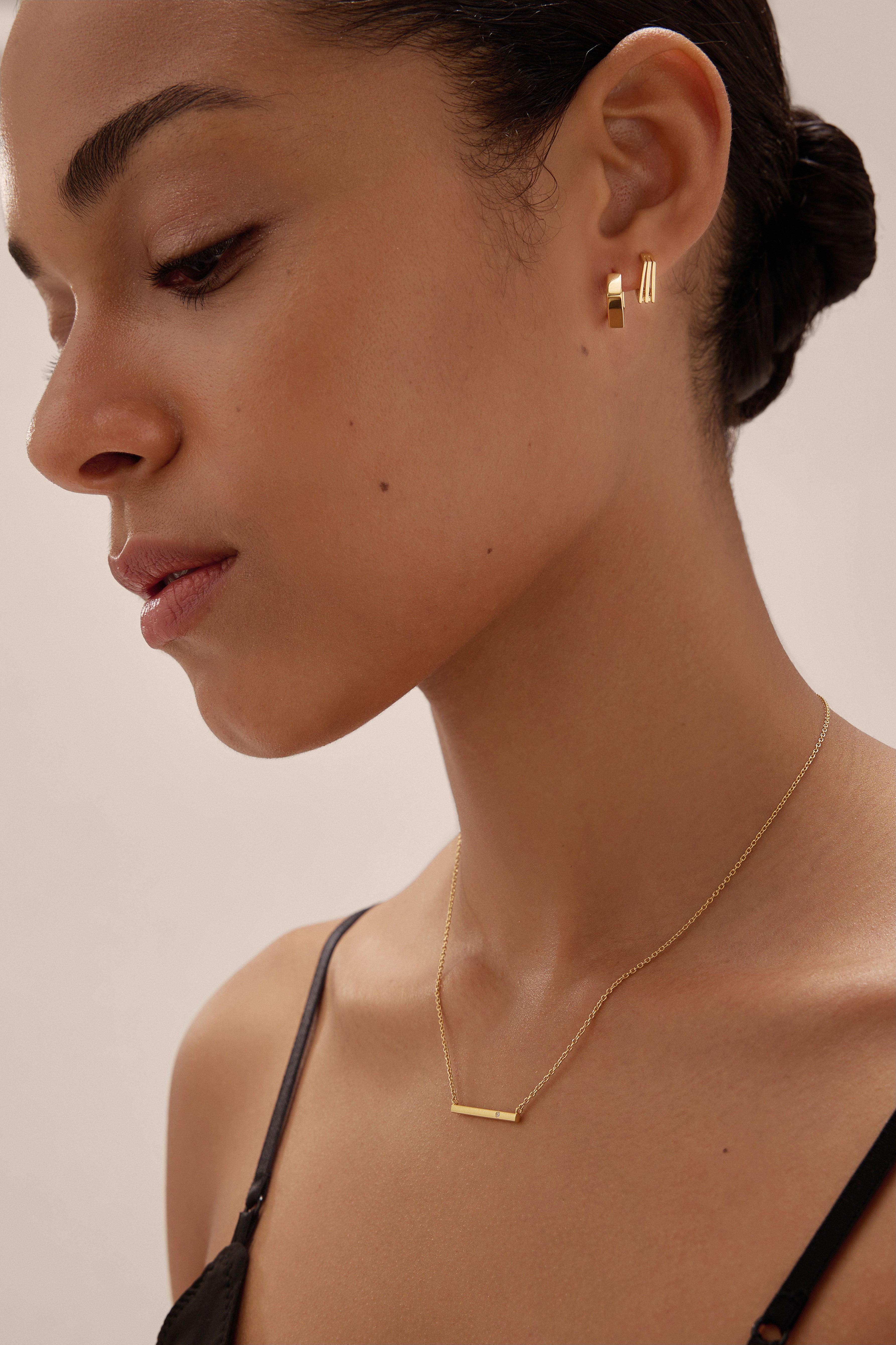 A woman with smooth skin and dark hair tied back wears the Juliet Angular Huggies in Gold, a delicate gold bar necklace, and a black strap top, photographed in soft lighting against a neutral background.