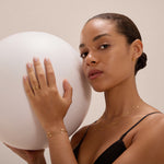 A woman with slicked-back hair in a black top holds a white balloon, wearing gold jewelry including the Juliet Angular Huggies in Gold. She gazes confidently at the camera against a neutral background.