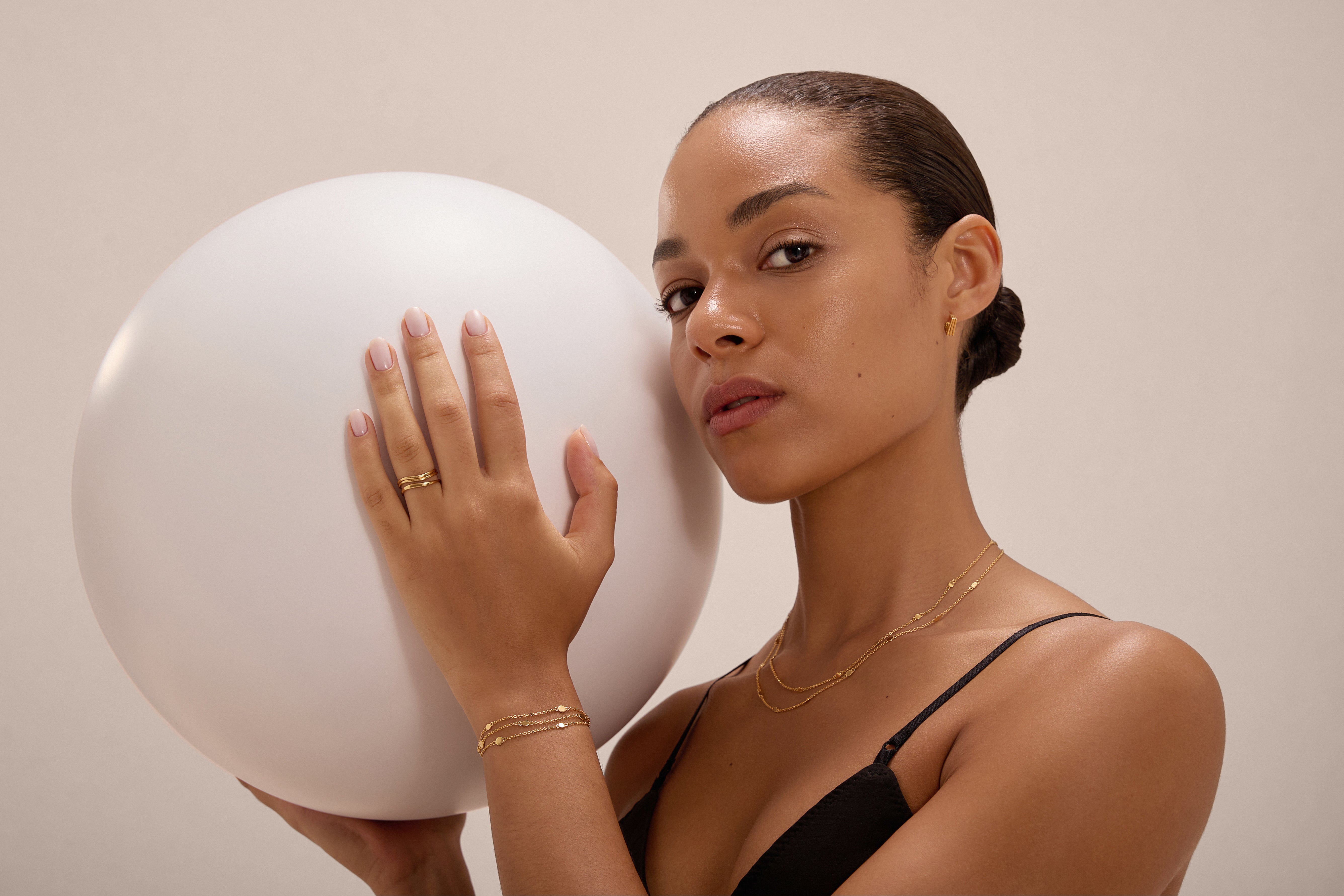 A woman with slicked-back hair in a black top holds a white balloon, wearing gold jewelry including the Juliet Angular Huggies in Gold. She gazes confidently at the camera against a neutral background.