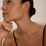 A woman with smooth skin and slicked-back hair wears a black top, the Juliet Angular Huggies in Gold, and a delicate gold necklace with green gemstones. She gazes upward thoughtfully, her hand resting lightly on her chin.