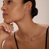 A woman with smooth skin and slicked-back hair wears a black top, the Juliet Angular Huggies in Gold, and a delicate gold necklace with green gemstones. She gazes upward thoughtfully, her hand resting lightly on her chin.
