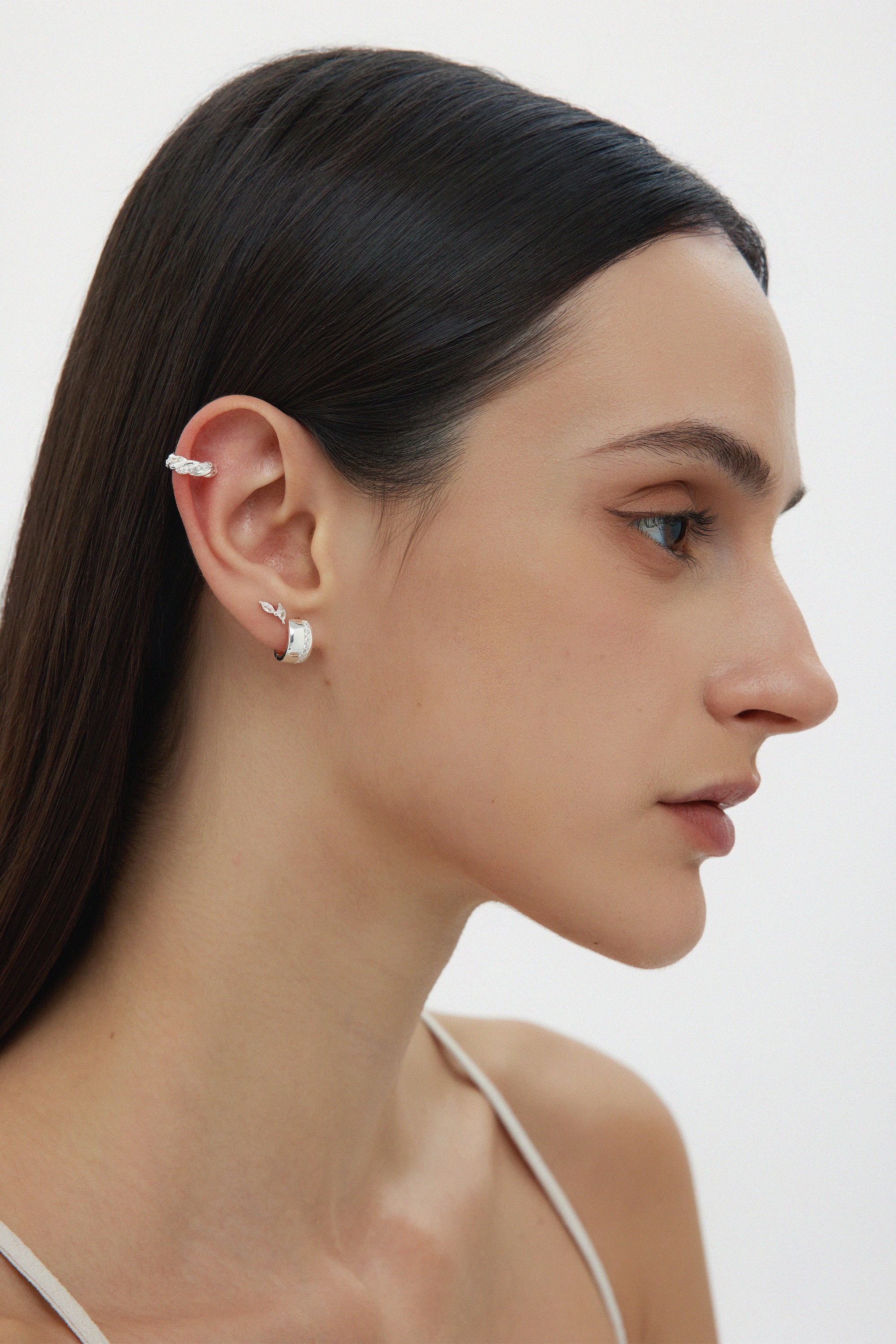 A woman with straight dark hair, in a white thin-strap top, is shown in profile wearing sterling silver ear cuffs and Juni Pavé Chunky Hoop Earrings in Silver on her left ear, against a plain white background.
