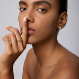 A woman with smooth skin and neat brows poses against a plain light background, finger to her lips, wearing Juni Pavé Chunky Huggie Earrings in Gold and rings, gazing directly at the camera.