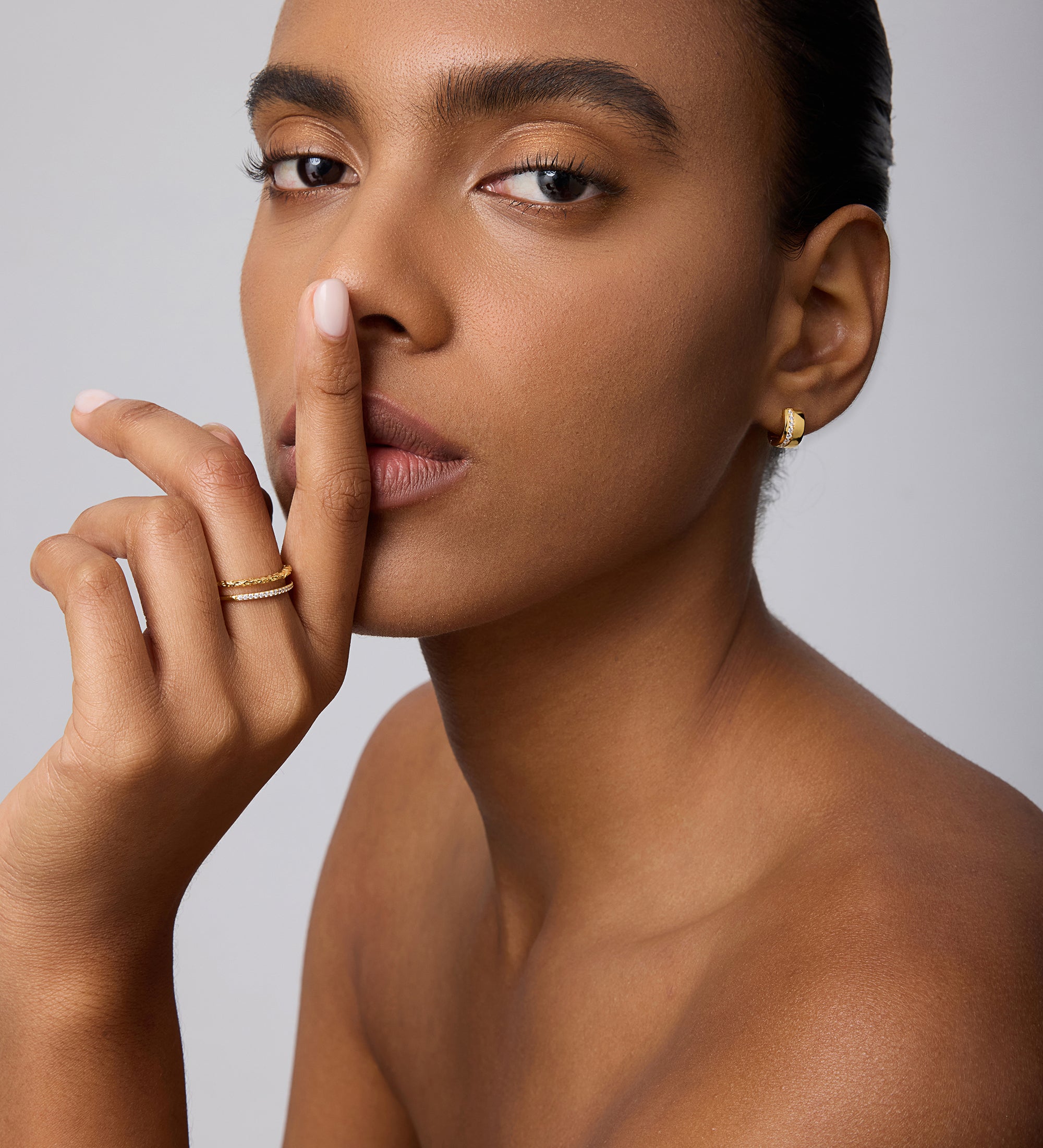 A woman with smooth skin and neat brows poses against a plain light background, finger to her lips, wearing Juni Pavé Chunky Huggie Earrings in Gold and rings, gazing directly at the camera.