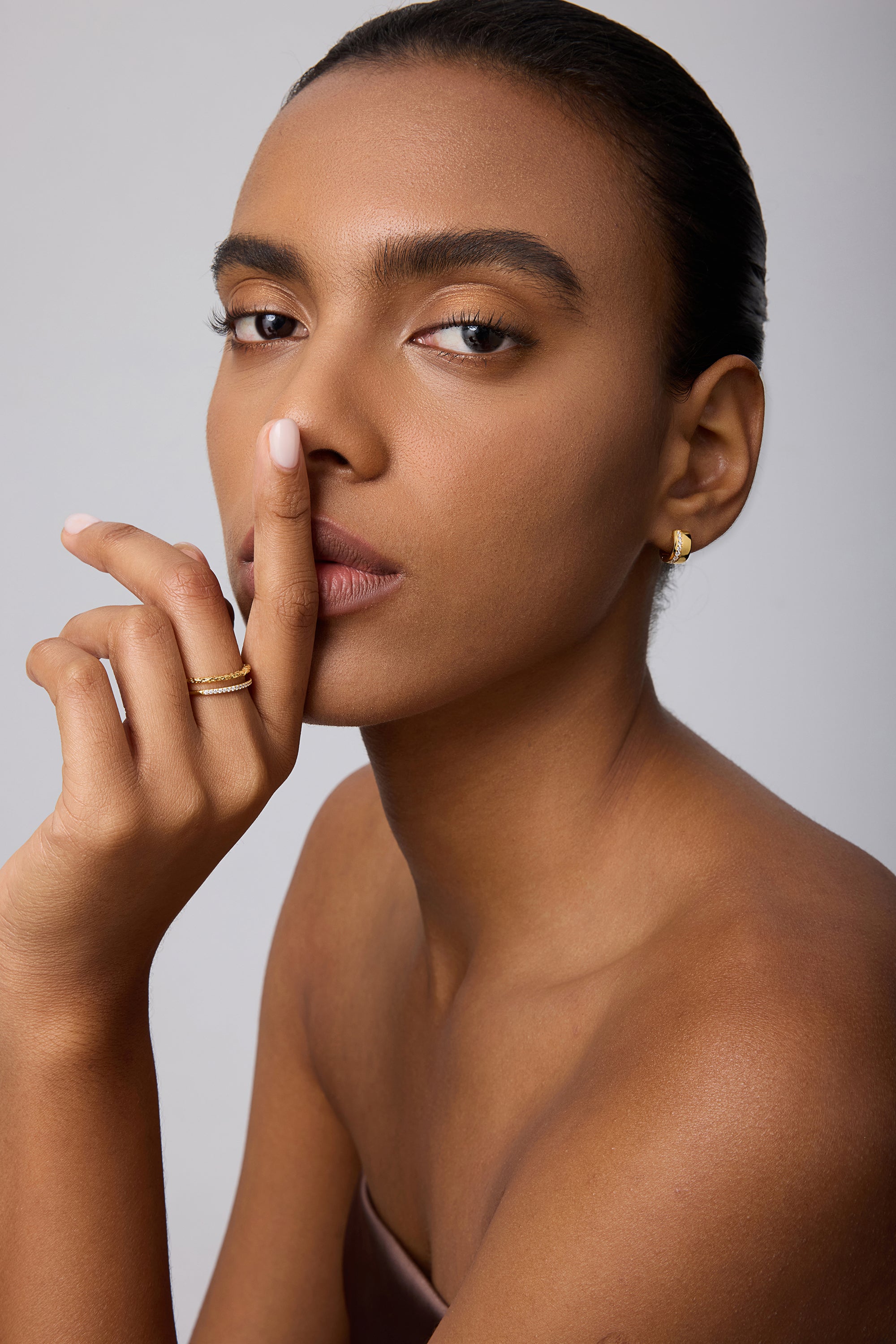 A woman with smooth skin and neat brows poses against a plain light background, finger to her lips, wearing Juni Pavé Chunky Huggie Earrings in Gold and rings, gazing directly at the camera.