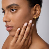 A woman with smooth skin and manicured brows wears the Juni Pavé Chunky Huggie Earrings in Gold, gently touching her face to showcase her jewelry against a neutral background.