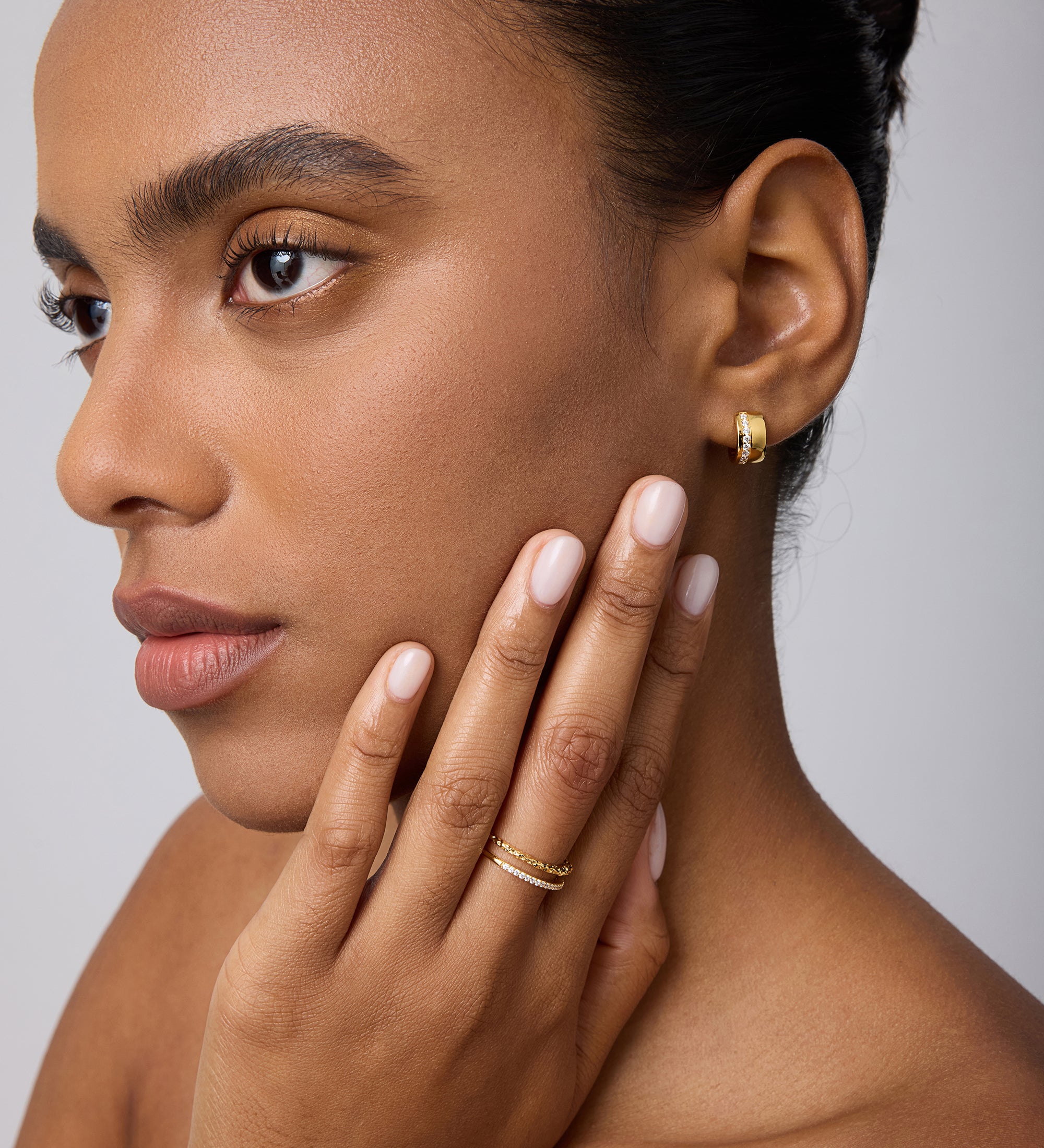 A woman with smooth skin and manicured brows wears the Juni Pavé Chunky Huggie Earrings in Gold, gently touching her face to showcase her jewelry against a neutral background.