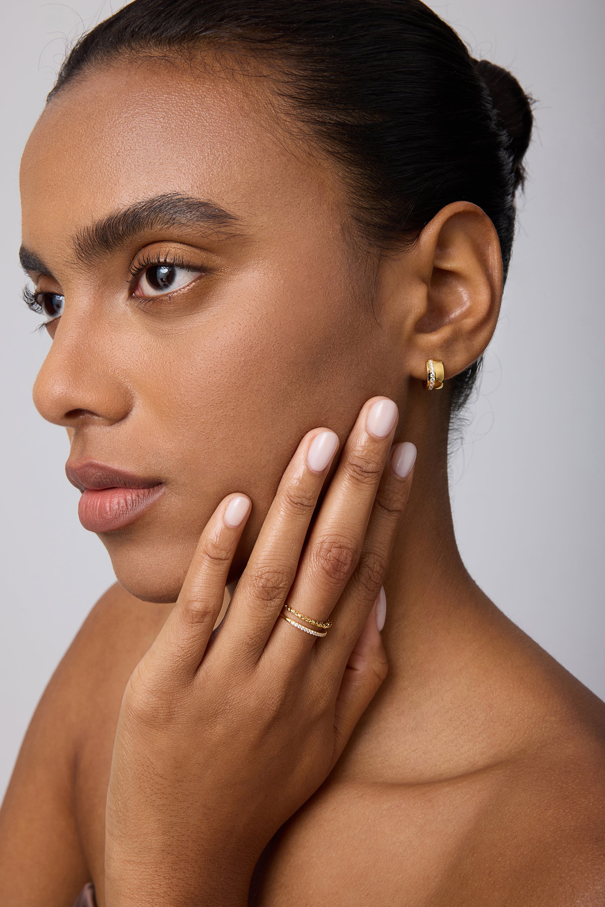 A woman with smooth skin and manicured brows wears the Juni Pavé Chunky Huggie Earrings in Gold, gently touching her face to showcase her jewelry against a neutral background.