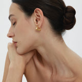 A woman with dark hair in a neat bun poses in profile, eyes closed and chin on her hand. She wears the Juno Ridged Chunky Hoops in Gold and a strapless top against a plain, light background.