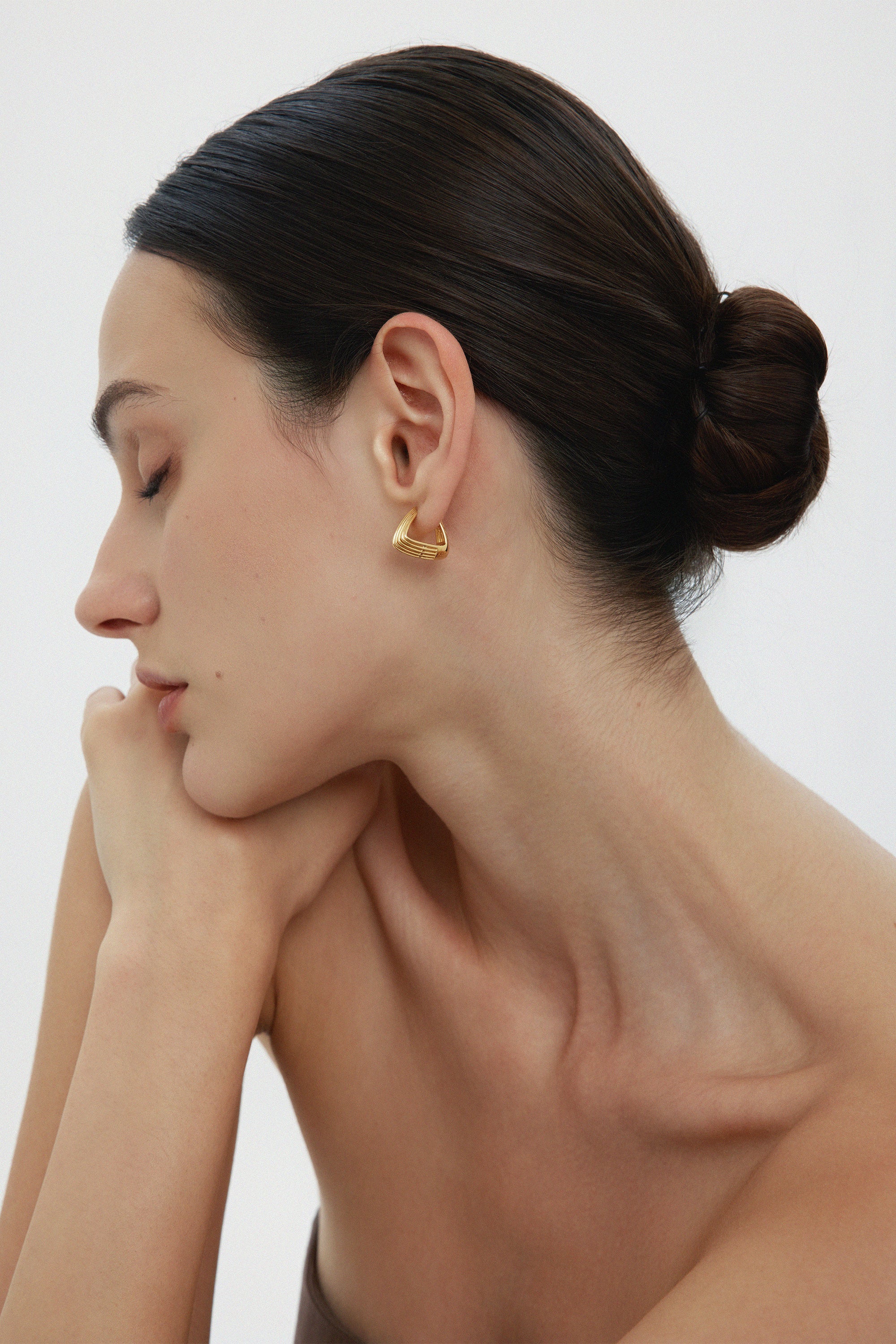 A woman with dark hair in a neat bun poses in profile, eyes closed and chin on her hand. She wears the Juno Ridged Chunky Hoops in Gold and a strapless top against a plain, light background.