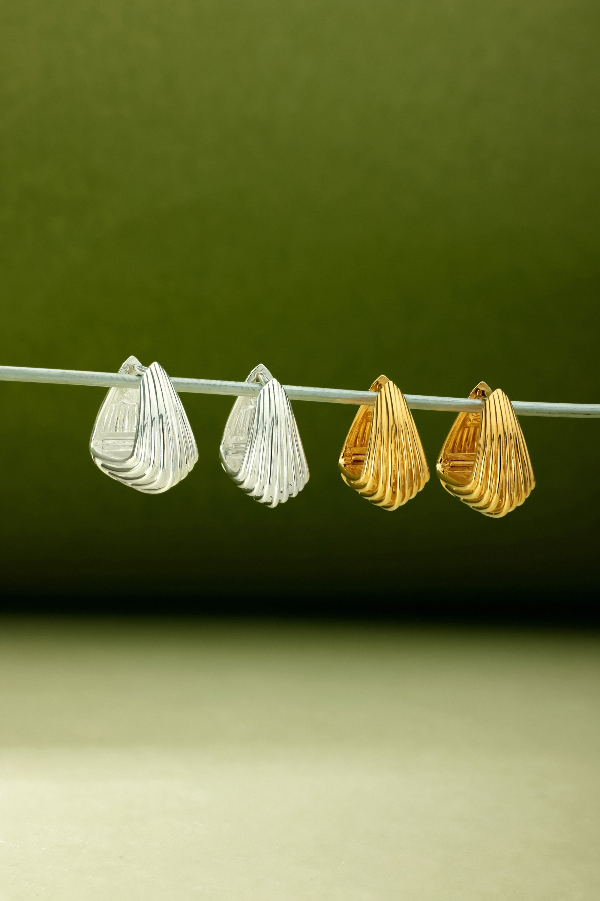 The Juno Ridged Chunky Hoops in Gold hang side by side with a silver pair on a thin rod against a green gradient backdrop, highlighting their modern textured design and elegant chunky gold finish.