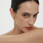A woman with slicked-back dark hair and bare shoulders gazes at the camera, wearing Juno Ridged Chunky Hoops in Gold and resting her arm across her chest against a plain, light background.