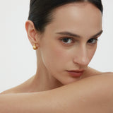 A woman with slicked-back dark hair and bare shoulders gazes at the camera, wearing Juno Ridged Chunky Hoops in Gold and resting her arm across her chest against a plain, light background.