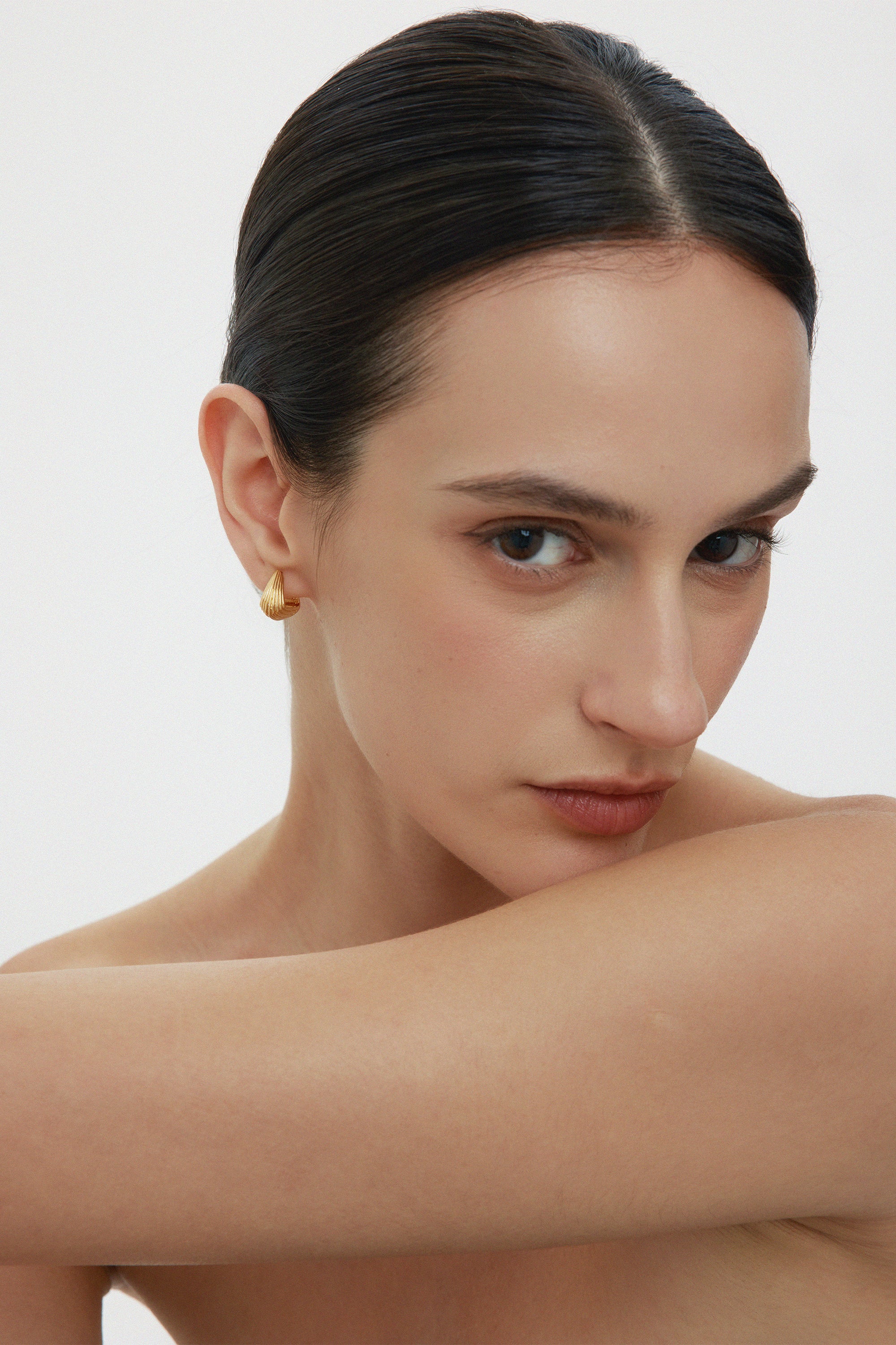 A woman with slicked-back dark hair and bare shoulders gazes at the camera, wearing Juno Ridged Chunky Hoops in Gold and resting her arm across her chest against a plain, light background.