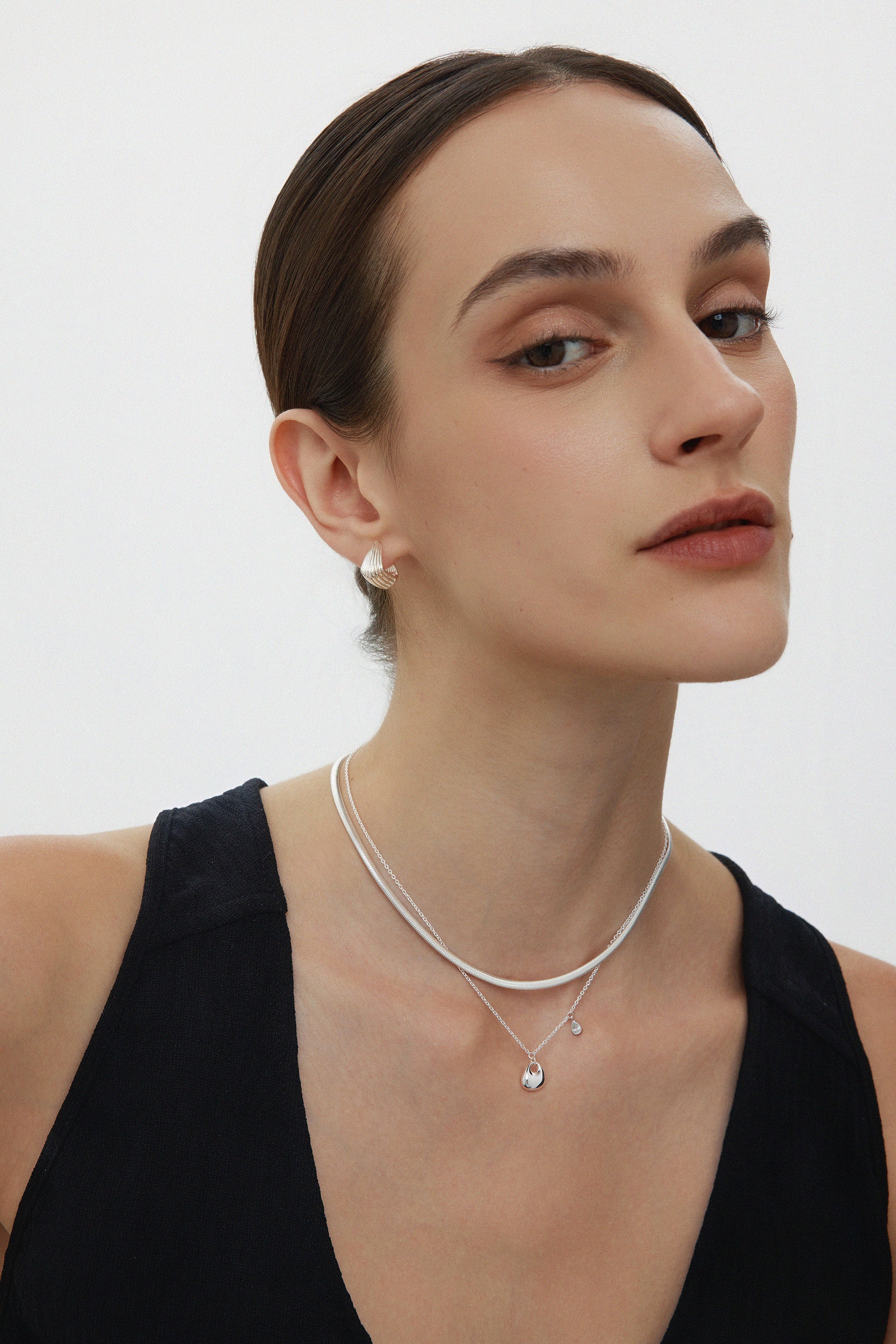 A woman with slicked-back brown hair wears a sleeveless black top and Juno Ridged Chunky Hoops in Silver, looking confidently at the camera against a plain white background.