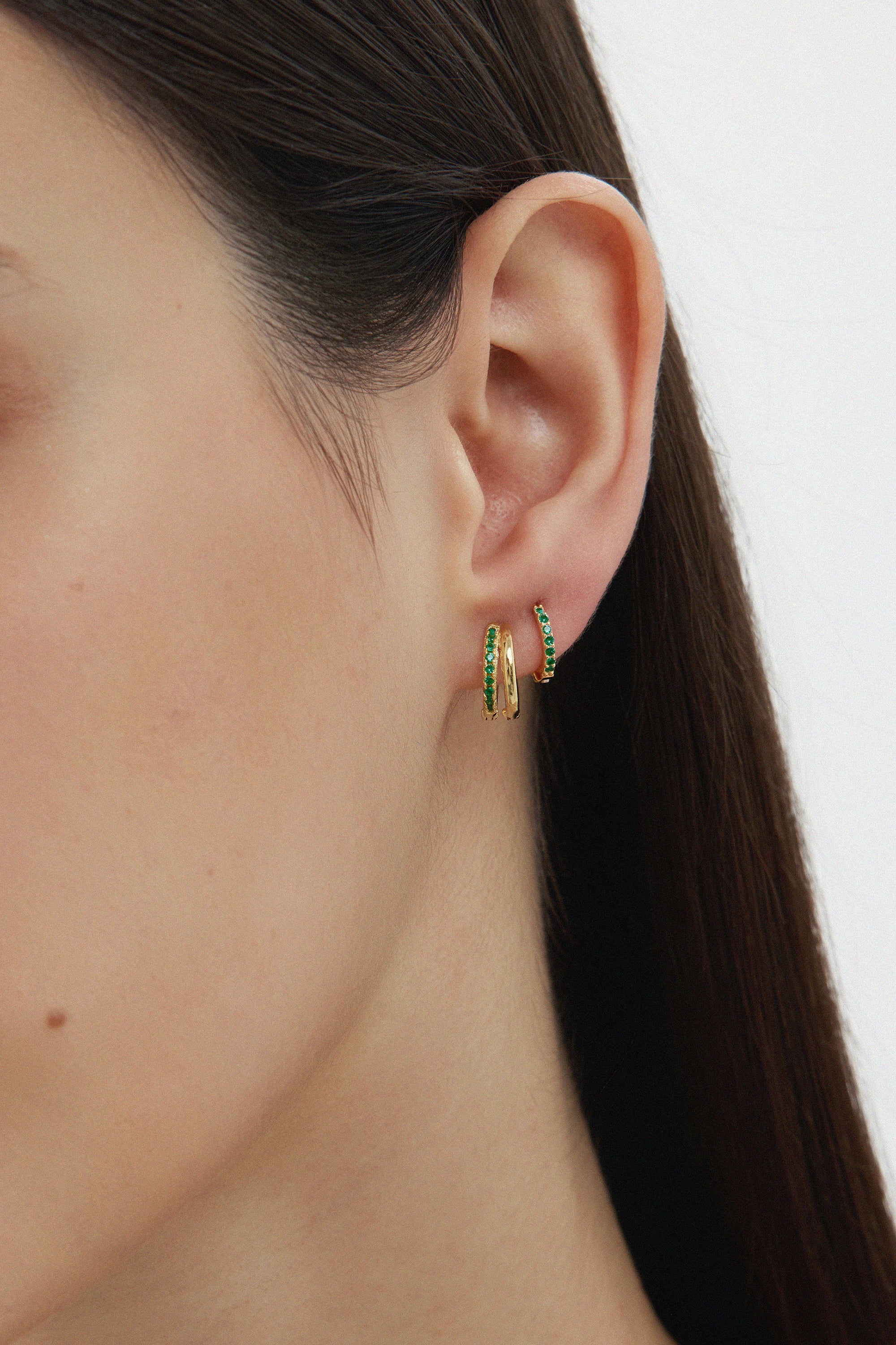 Close-up of a womans ear with two gold hoops—one plain, one Kiki Crystal Huggie Earring in Gold/Green. She has straight, dark hair and smooth skin, set against a light background.