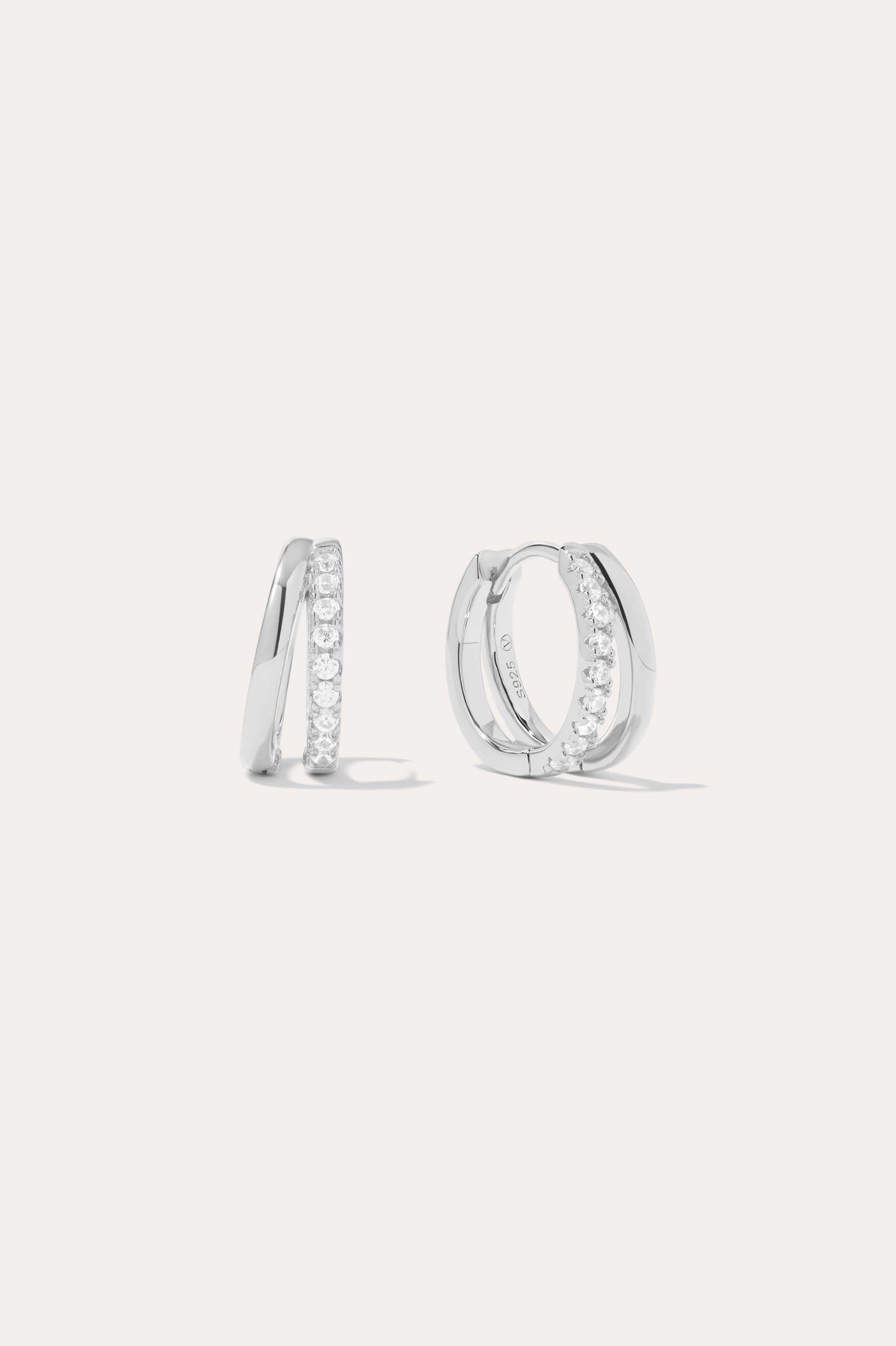 The Kira Crystal Double Hoop Huggies in Sterling Silver are shown, each featuring a row of sparkling round crystals along the front edge. The background is plain and light-colored.