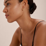 A woman with smooth, medium-brown skin and a low bun wears a black top, the Lila Medium Round Dome Hoop Earrings in Gold, and a delicate gold necklace. She faces left against a softly lit, neutral background.