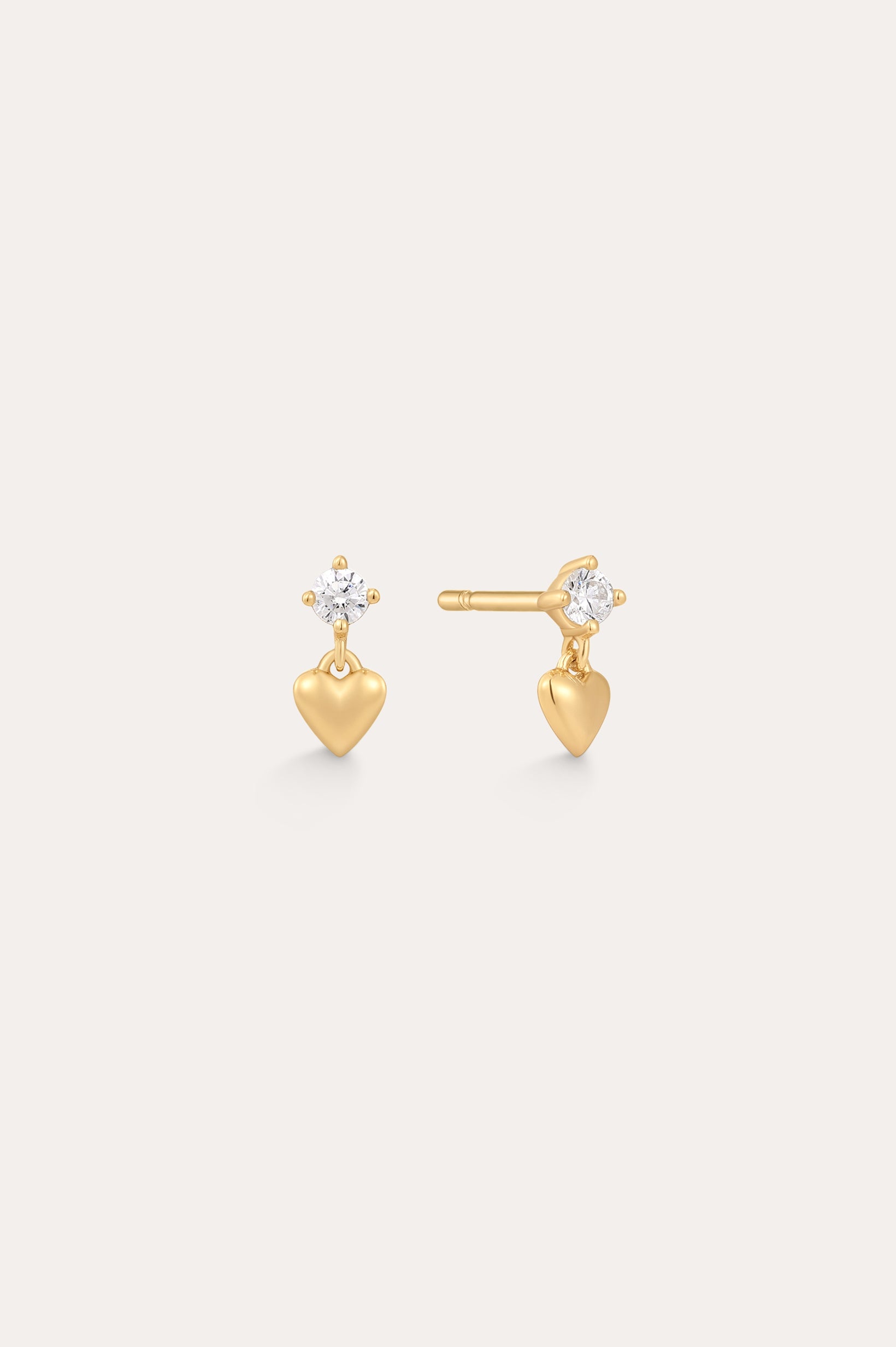 The Lilian Crystal Heart Studs Earrings in Gold feature a small gold heart charm dangling below a sparkling round crystal, displayed on a plain light background.