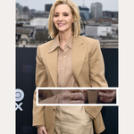 A woman with short blonde hair smiles outdoors in a city, wearing a beige blazer over a matching blouse and the Ayla Link Ring in Gold. Text above reads SPOTTED ON: and below, LISA KUDROW.