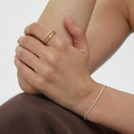 A close-up of a person’s hands resting on their knee, wearing a gold ring and the Lumi Tennis Bracelet in Gold with cubic zirconia, against soft brown fabric and a neutral background.