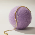 The Lumi Tennis Bracelet in Gold, adorned with sparkling cubic zirconia gemstones, is elegantly draped over a large fuzzy purple ball against a simple light background.