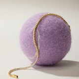 The Lumi Tennis Bracelet in Gold, adorned with sparkling cubic zirconia gemstones, is elegantly draped over a large fuzzy purple ball against a simple light background.