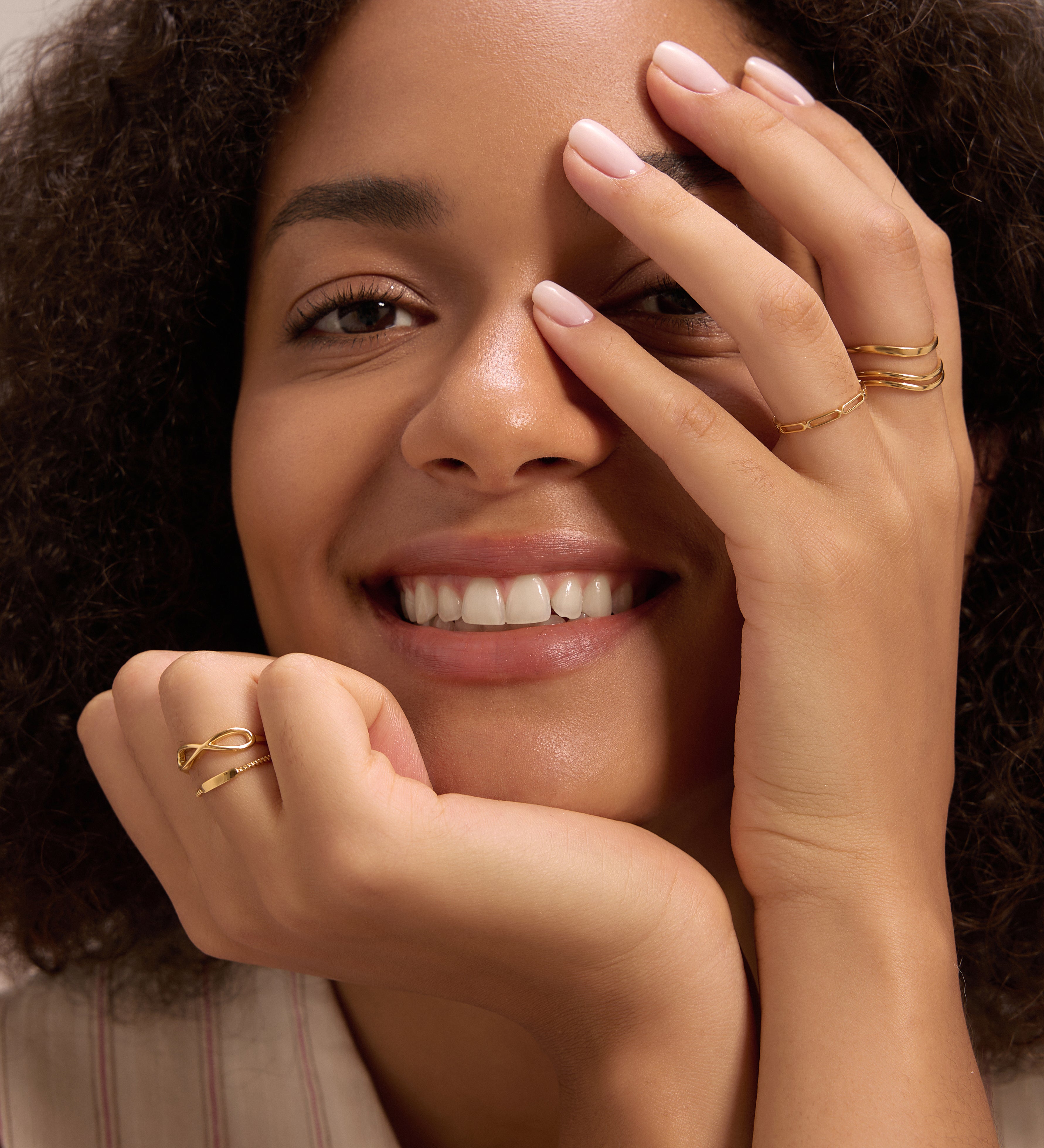 A smiling woman with curly hair rests her chin on one hand, displaying the Lya Horizontal Bar Stacking Ring in Gold on the other. She wears light pink nail polish, showcasing an effortless and elegant style.