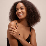 A woman with curly hair wears a black tank top and smiles while looking to the side. She accessorizes with gold jewelry, including the Lya Horizontal Bar Stacking Ring in Gold, along with other rings, bracelets, and layered necklaces.