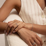 A woman wears multiple stackable rings, including the Lya Horizontal Bar Stacking Ring in Silver, with manicured nails and a delicate bracelet, seated in a sleeveless, light-striped dress on a textured chair.