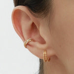 A close-up of a persons ear wearing Mae Twist Double Huggie Earrings in Gold—featuring a textured rope hoop on the lobe and a gold huggie on the cartilage. The background is plain, and hair is tucked behind the ear.