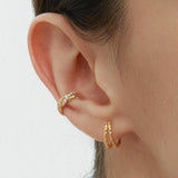 A close-up of a persons ear wearing Mae Twist Double Huggie Earrings in Gold—featuring a textured rope hoop on the lobe and a gold huggie on the cartilage. The background is plain, and hair is tucked behind the ear.