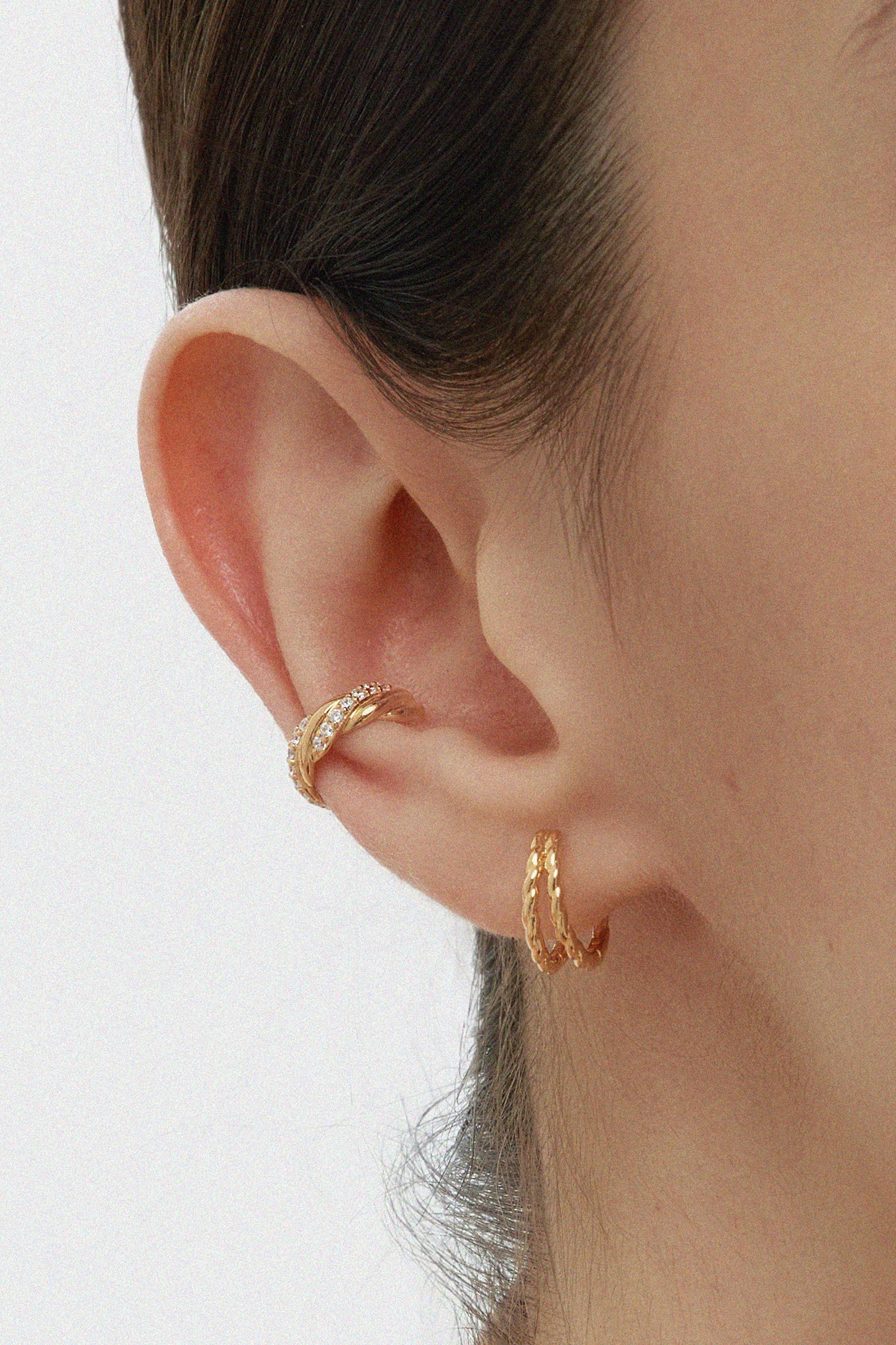 A close-up of a persons ear wearing Mae Twist Double Huggie Earrings in Gold—featuring a textured rope hoop on the lobe and a gold huggie on the cartilage. The background is plain, and hair is tucked behind the ear.