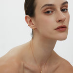 A woman with slicked-back dark hair wears the Mae Twist Double Huggie Earrings in Gold and a delicate gold necklace. Sporting natural makeup, she gazes confidently at the camera against a plain white background.