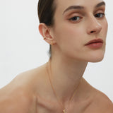 A woman with slicked-back dark hair wears the Mae Twist Double Huggie Earrings in Gold and a delicate gold necklace. Sporting natural makeup, she gazes confidently at the camera against a plain white background.