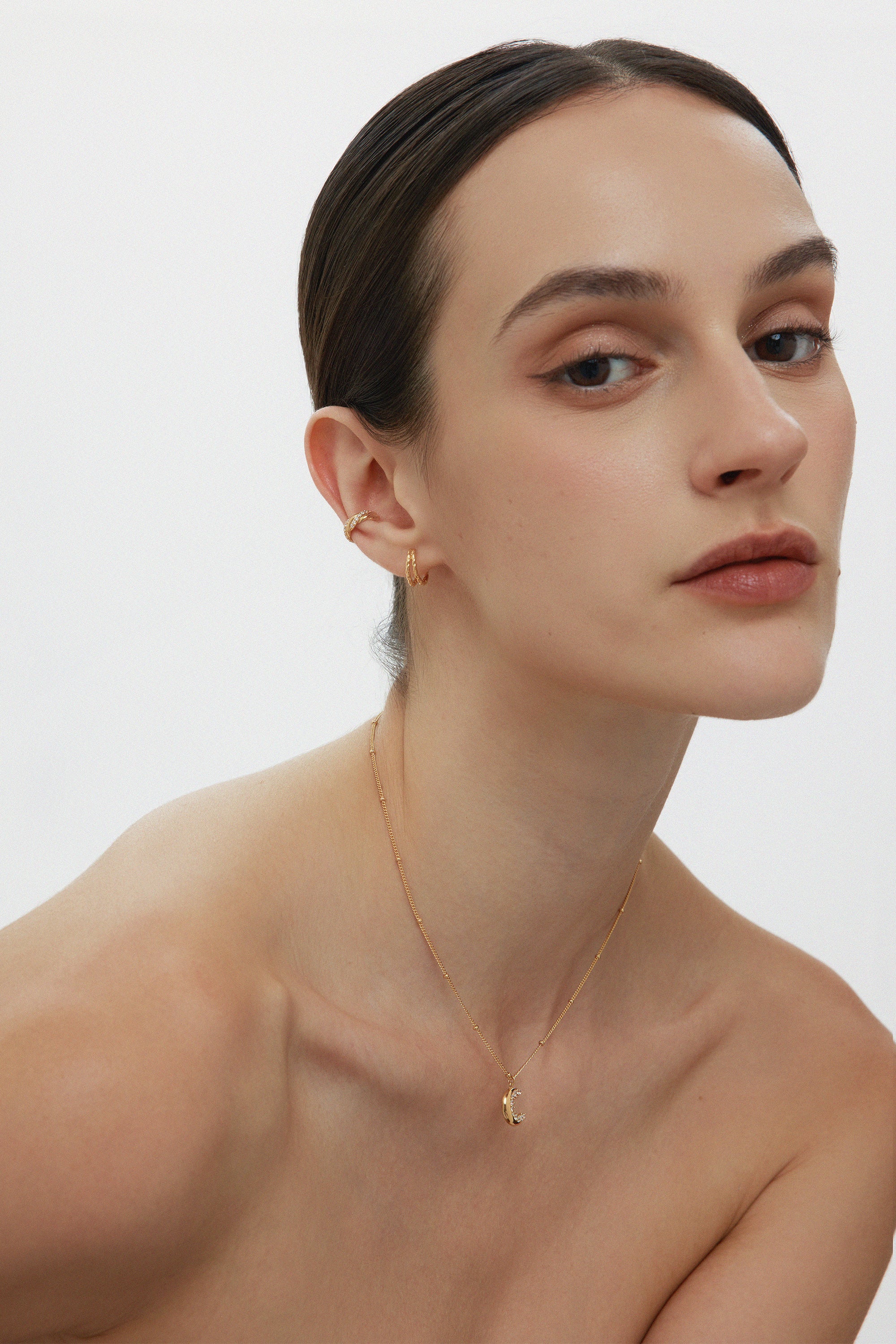 A woman with slicked-back dark hair wears the Mae Twist Double Huggie Earrings in Gold and a delicate gold necklace. Sporting natural makeup, she gazes confidently at the camera against a plain white background.