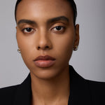 A woman with smooth brown skin and straight dark hair pulled back wears the Mia Two-tone Mixed Metal Hoops and a black blazer, gazing directly at the camera with a neutral expression against a plain background.