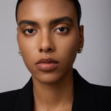 A woman with smooth brown skin and straight dark hair pulled back wears the Mia Two-tone Mixed Metal Hoops and a black blazer, gazing directly at the camera with a neutral expression against a plain background.