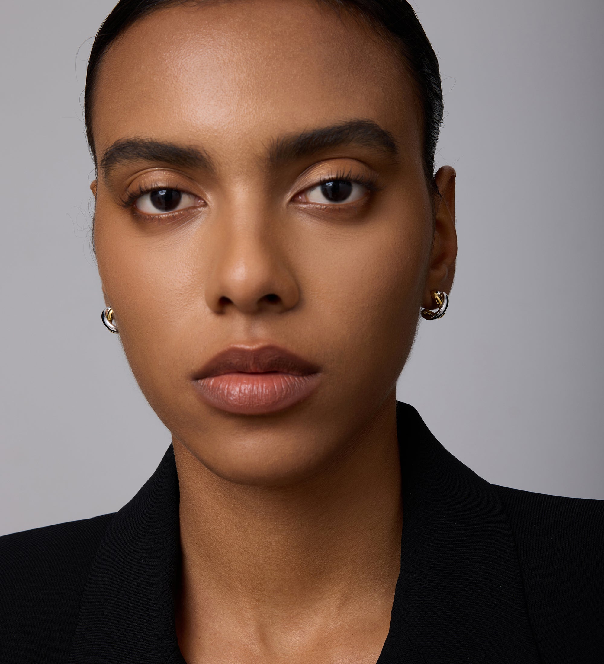 A woman with smooth brown skin and straight dark hair pulled back wears the Mia Two-tone Mixed Metal Hoops and a black blazer, gazing directly at the camera with a neutral expression against a plain background.