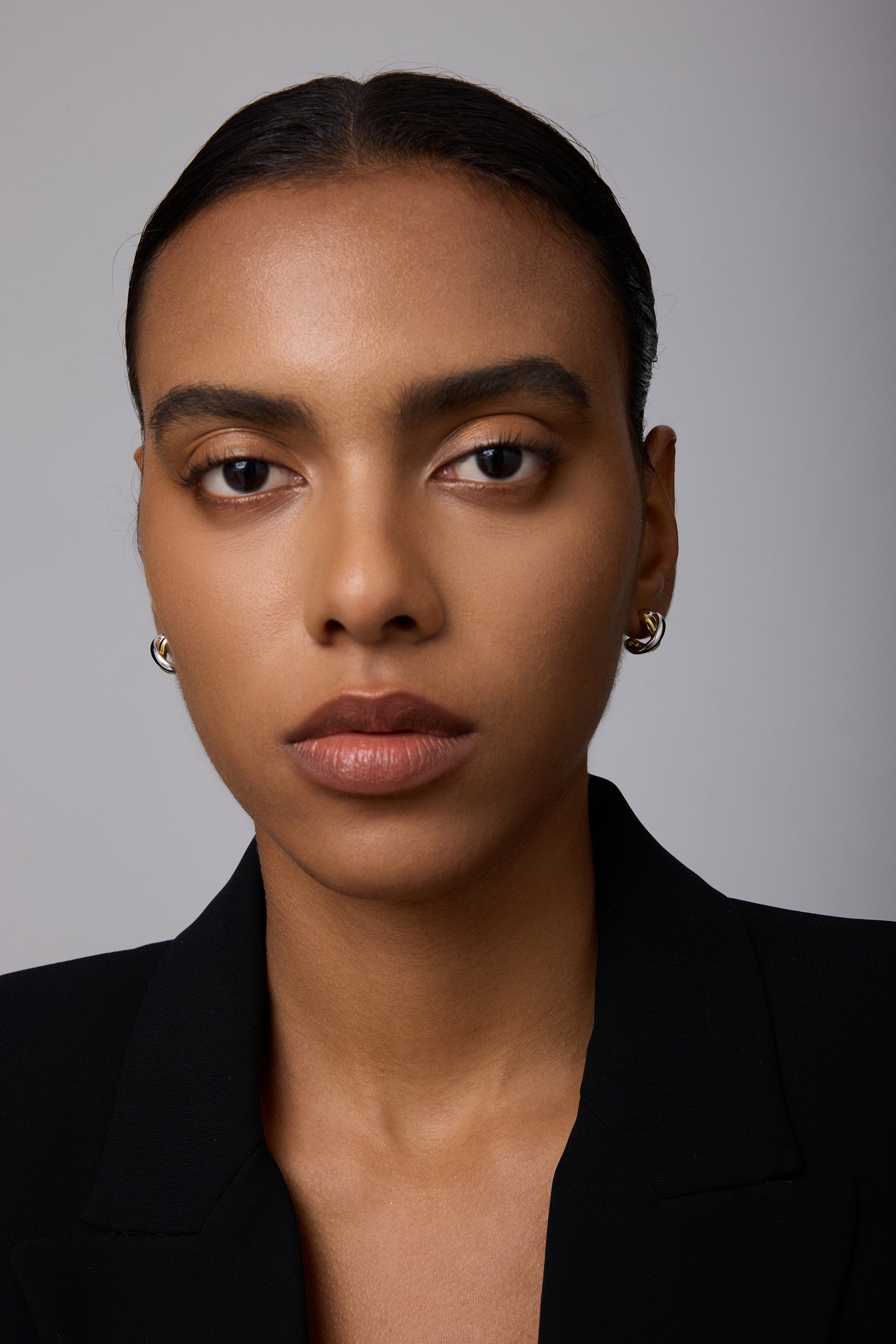 A woman with smooth brown skin and straight dark hair pulled back wears the Mia Two-tone Mixed Metal Hoops and a black blazer, gazing directly at the camera with a neutral expression against a plain background.