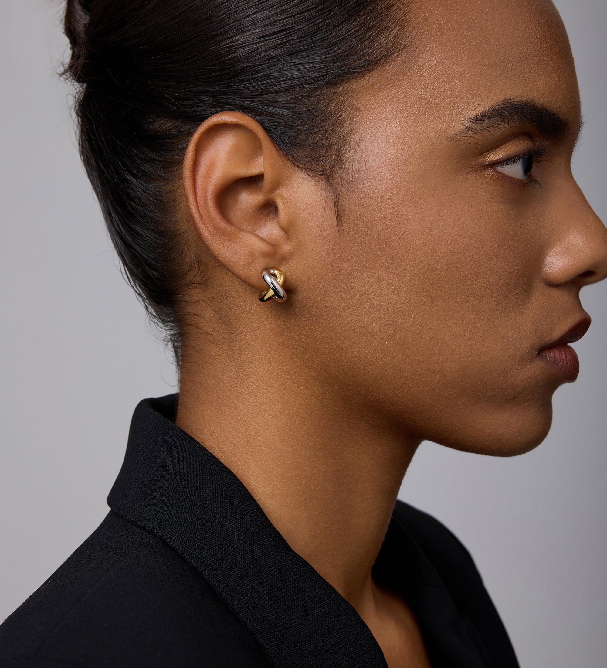 A woman with dark hair in a neat bun wears a black blazer and showcases the Mia Two-tone Mixed Metal Hoops. She appears in profile against a plain, light gray background.