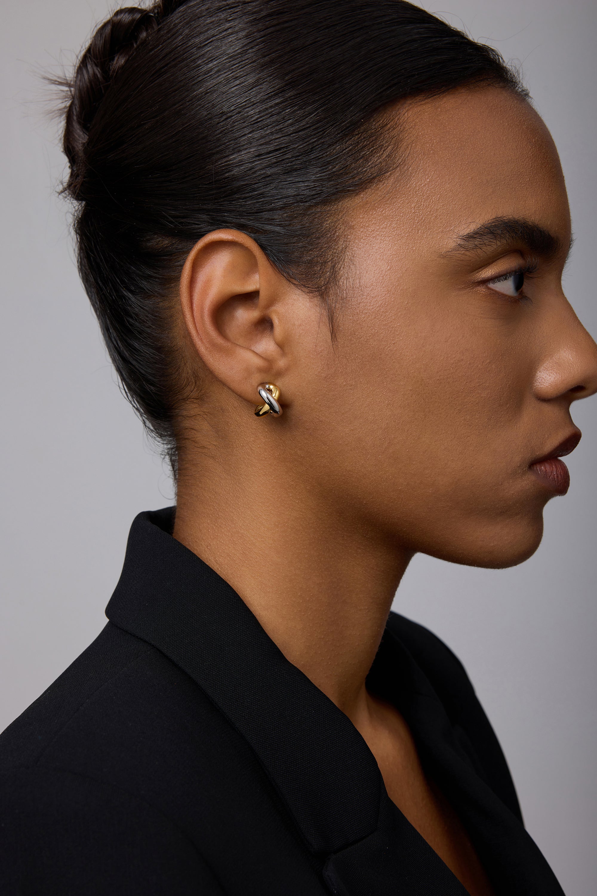 A woman with dark hair in a neat bun wears a black blazer and showcases the Mia Two-tone Mixed Metal Hoops. She appears in profile against a plain, light gray background.